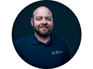 Porträt eines lächelnden Mannes mit kurzgeschnittenem Bart und Glatze, der ein dunkelblaues Poloshirt mit der Aufschrift „TÜV Rheinland FSP“ trägt. Der Hintergrund ist dunkelblau und kreisförmig.