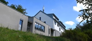 Modernes, mehrstöckiges Gebäude mit grauer und weißer Fassade auf einem begrünten Hang, im Hintergrund Bäume und blauer Himmel mit wenigen Wolken, auf dem Dach befindet sich eine Mobilfunkantenne.